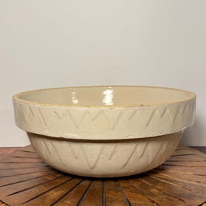 May include: A cream-colored ceramic mixing bowl with a decorative zigzag pattern around the rim. The bowl has a wide, shallow shape and a slightly textured surface. This vintage bowl is perfect for baking and cooking.