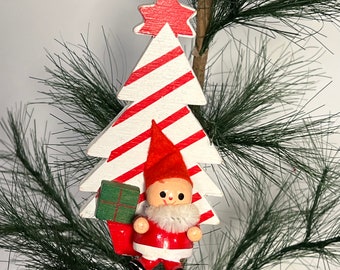 May include: A wooden Christmas tree ornament with a red and white striped tree and a small gnome holding a green gift box. The gnome is wearing a red hat, red coat, and white beard.