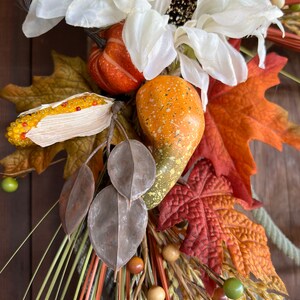 May include: A fall-themed wreath with white flowers, orange gourds, brown leaves, and wheat stalks. The wreath is decorated with small wooden beads and green berries.