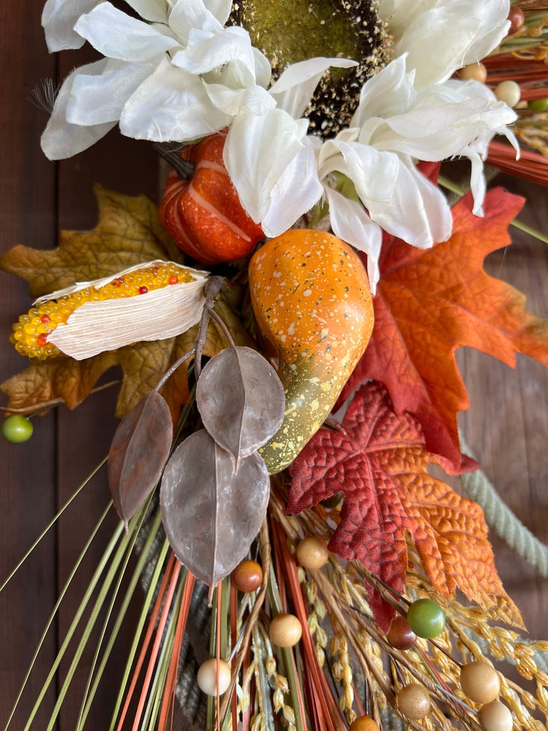 May include: A fall-themed wreath with white flowers, orange gourds, brown leaves, and wheat stalks. The wreath is decorated with small wooden beads and green berries.