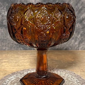 May include: A brown glass candy dish with a pedestal base and a geometric pattern. The dish has a scalloped edge and a hexagonal base.