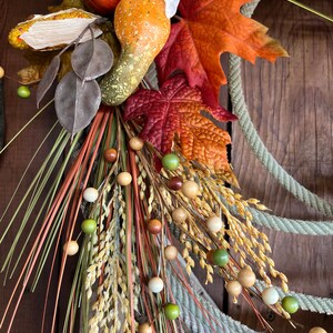 May include: A fall-themed wreath with a rope base, featuring a variety of autumnal elements, including gourds, leaves, and wheat stalks. The wreath is decorated with small wooden beads in shades of brown, beige, and green.