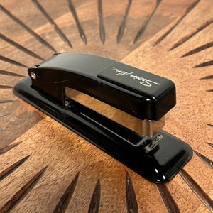 May include: A black metal stapler with a silver staple remover and the word "Swingline" printed on the top. The stapler is sitting on a wooden surface.