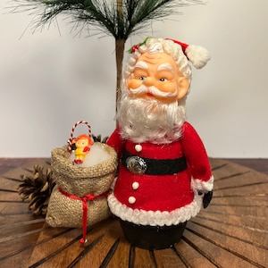 May include: A vintage Santa Claus figurine with a white beard and red suit, holding a burlap sack with a small doll inside. The figurine is standing on a wooden surface.