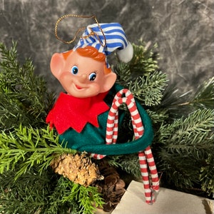 May include: A Christmas elf ornament with a red and green outfit, a blue and white striped hat, and a candy cane. The elf is holding a candy cane in its hand.