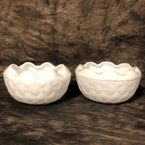 May include: Two white frosted glass bowls with a diamond pattern and scalloped edges. The bowls are empty and sitting on a brown textured surface.
