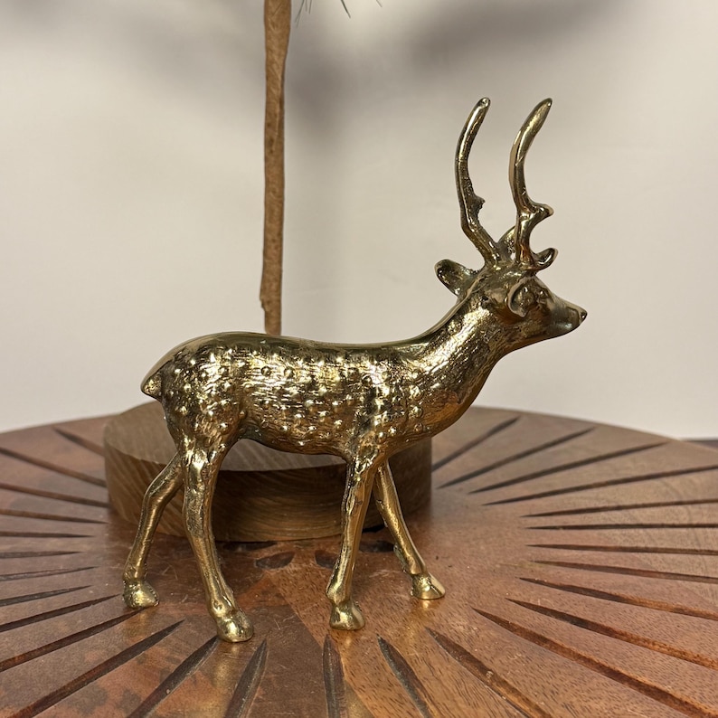 May include: A golden-colored deer figurine with detailed antlers and a textured body. The decorative sculpture is standing on a wooden surface, showcasing a classic and elegant design. The deer is a symbol of nature and grace.