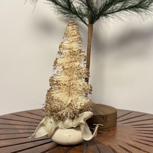May include: A small, decorative Christmas tree with a cream-colored, bristled texture and gold accents. The tree sits atop a small, white ceramic pot wrapped with a burlap ribbon. A small, green pine branch is in the background.