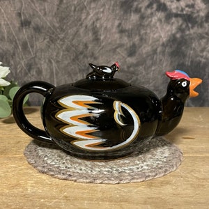 May include: Black ceramic teapot shaped like a chicken with white and orange painted details. The teapot has a small chicken figurine on the lid.