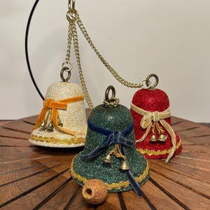 May include: Three decorative bell ornaments in cream, green, and red with gold glitter. Each bell has a velvet ribbon and small gold bells. They hang from a gold chain, perfect for holiday decor.