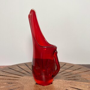 May include: A vibrant red glass vase with a unique, elongated shape and a small handle. The vase has a smooth, glossy finish and stands on a wooden surface. The design is reminiscent of mid-century modern decor.