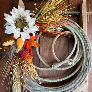 May include: A rustic fall wreath with a white sunflower, dried grasses, and small brown and green berries. The wreath is hanging on a weathered wooden fence with a coiled rope.