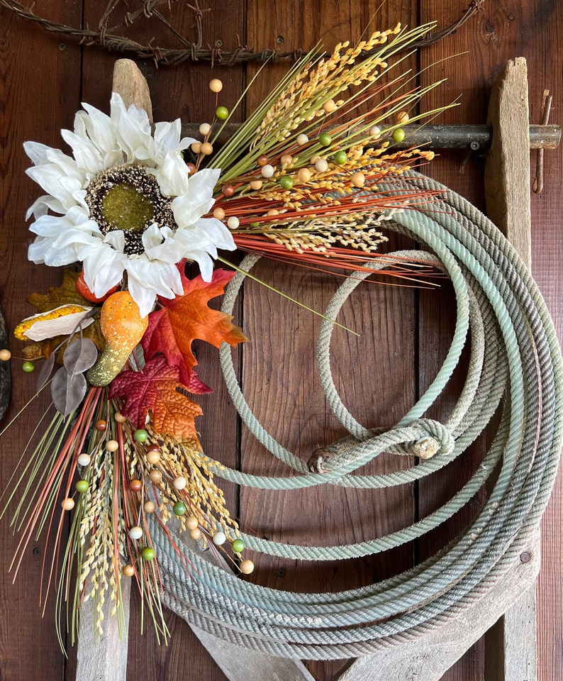May include: A rustic fall wreath with a white sunflower, dried grasses, and small brown and green berries. The wreath is hanging on a weathered wooden fence with a coiled rope.