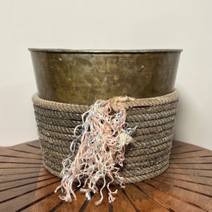 May include: A gold metal planter with a rope trim and a pink and blue tassel. The planter is sitting on a wooden surface.