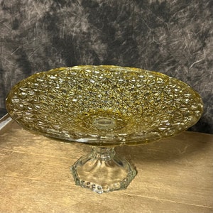 May include: A clear glass pedestal cake stand with a gold-toned, diamond-patterned bowl. The stand has a scalloped base.
