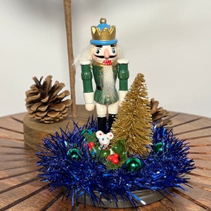 May include: A wooden nutcracker figurine wearing a green and white uniform with a blue and gold crown stands in front of a small gold pine tree. The nutcracker is surrounded by blue tinsel, green holly, and red berries.