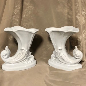 May include: A pair of white ceramic vases with a decorative, ornate, scroll-like design. The vases are shaped like a horn or trumpet.