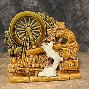 May include: A ceramic wall plaque featuring a spinning wheel, a cat, and a dog. The plaque is brown and green with white accents. The spinning wheel is made of wood and has a brown wooden frame. The cat is sitting on the spinning wheel and is looking at the dog. The dog is standing on the ground and is looking at the cat. The plaque has the letters "R.S." on the bottom right corner.
