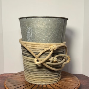 May include: A galvanized metal bucket with a rope-wrapped base and handle. The bucket has a silver finish and a cylindrical shape. The rope is beige and gray, adding a rustic touch. The bucket sits on a wooden surface.