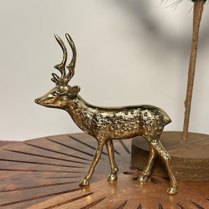 May include: A golden brass deer figurine with detailed antlers and a textured body. The deer is standing on a wooden surface, showcasing its elegant form. This decorative piece adds a touch of nature-inspired charm to any space.