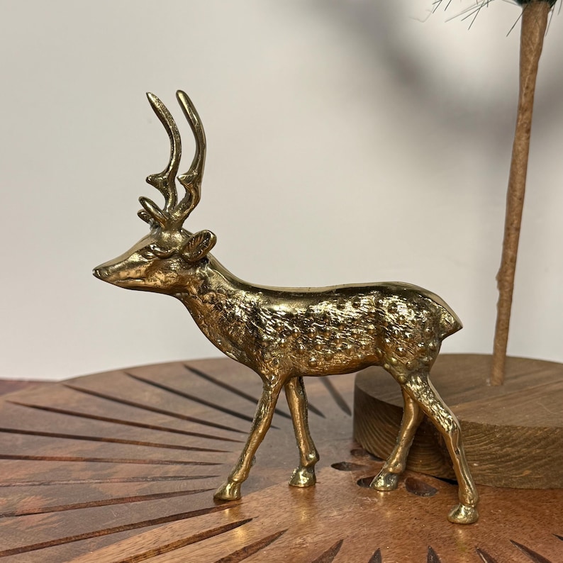 May include: A golden brass deer figurine with detailed antlers and a textured body. The deer is standing on a wooden surface, showcasing its elegant form. This decorative piece adds a touch of nature-inspired charm to any space.