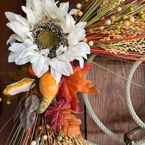 May include: A white sunflower with a brown center, surrounded by fall foliage, including red and orange leaves, wheat stalks, and brown and green beads. The wreath is attached to a rope.