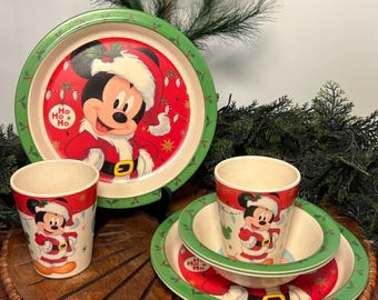 May include: A Christmas-themed Mickey Mouse dinnerware set. The set includes plates, bowls, and cups, all featuring Mickey in a Santa suit. The plates and bowls have a red background with a green border. The cups and bowls have a beige interior.