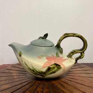 May include: A vintage ceramic teapot with a floral design. The teapot is light blue and white with a pink flower and green leaves. The handle is shaped like a branch and the lid has a small bird finial.