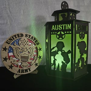 May include: Wooden United States Army themed decor. A round wooden piece with the words "United States Army" and a patriotic design. A black lantern with the name "Austin" and Army silhouettes is also present.
