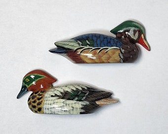 May include: Two hand-painted wooden duck figurines. The larger duck is blue, green, and brown with a red beak. The smaller duck is green, brown, and white with a red beak.