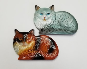 May include: Two ceramic cat figurines, one is a light blue and white cat, the other is a brown, black, and white cat.