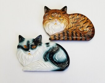 May include: Two ceramic cat figurines, one with black and white fur and the other with brown and orange fur. Both cats are lying down with their heads turned to the side.