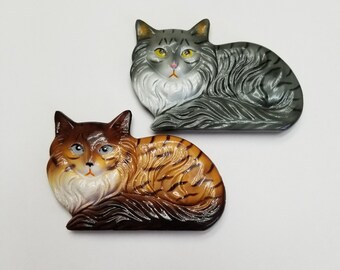 May include: Two ceramic cat figurines, one gray and one brown with black stripes. Both cats are lying down with their heads turned to the side.