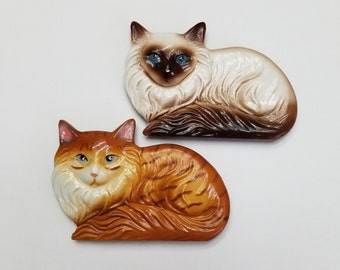 May include: Two ceramic cat figurines, one white with brown markings and blue eyes, the other orange with brown stripes and green eyes.
