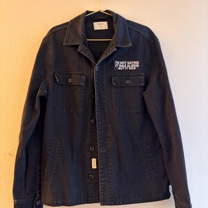 May include: A black button-down jacket with a collar, long sleeves, and two chest pockets. A rectangular patch on the left chest features the white text: I'M NOT SAYING IT WAS ALIENS BUT IT WAS. The jacket has a central button closure and a small label.