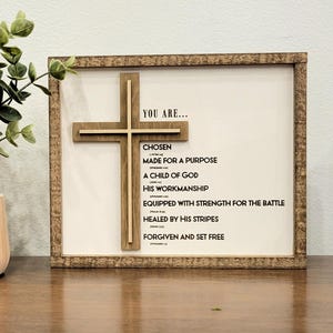 May include: A wooden sign with a cross design and the text "YOU ARE... CHOSEN MADE FOR A PURPOSE A CHILD OF GOD HIS WORKMANSHIP EQUIPPED WITH STRENGTH FOR THE BATTLE HEALED BY HIS STRIPES FORGIVEN AND SET FREE"