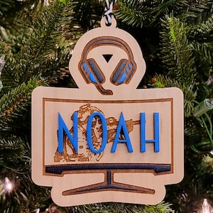 May include: Wooden Christmas ornament featuring a gamer theme. The ornament has a monitor shape with the name "NOAH" in blue letters. Above the monitor is a headset design. The ornament is hanging on a Christmas tree.