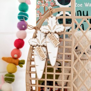 May include: Two white wooden carrot-shaped gift tags with the names "Sophie" and "Penelope" written on them. The tags are hanging from a brown wicker basket.