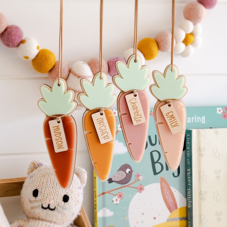 Puede incluir: Cuatro adornos de madera con forma de zanahoria con tapas verdes y etiquetas de nombre personalizadas. Los nombres en las etiquetas son Hudson, Archer, Charlotte y Emily. Las zanahorias est&aacute;n colgadas de una pared blanca.