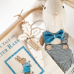 May include: A white canvas tote bag with the text "The Tale of Peter Rabbit" printed on the front. A wooden carrot-shaped tag with the name "Hudson" is attached to the bag. A plush bunny toy with a blue bow tie and striped overalls is sitting in the bag.