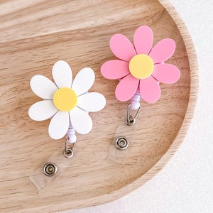 May include: Two daisy-shaped retractable badge holders. One is white with a yellow centre, the other is pink with a yellow centre. Both have a clear plastic strap and a metal clip for attaching to clothing.