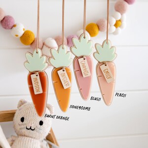 Puede incluir: Cuatro adornos de madera en forma de zanahoria con tapas verdes y etiquetas de nombre personalizadas. Las zanahorias son de color naranja quemado, miel, rubor y melocot&oacute;n.