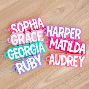 May include: A collection of colourful name tags in shades of pink, purple, and teal. Each tag features a different name in bold white lettering: Sophia, Grace, Georgia, Ruby, Harper, Matilda, and Audrey. Ideal for luggage or personal belongings.