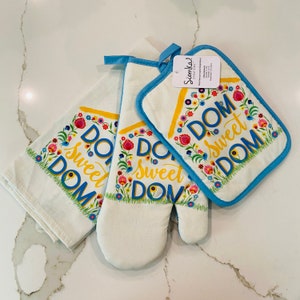 May include: A white kitchen set with the words "DOM Sweet DOM" in blue and yellow, surrounded by a floral design. The set includes a towel, oven mitt, and pot holder with blue trim.