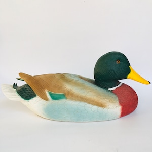 Puede incluir: Un señuelo de pato pintado con cabeza verde, pico amarillo y alas marrones y blancas. El pato tiene el pecho rojo y el vientre azul claro. El señuelo está diseñado para flotar en el agua y atraer a otros patos.