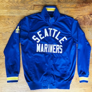 May include: A royal blue Seattle Mariners zip-up jacket with white lettering. The jacket features the team name across the chest and a yellow and white trim on the collar, cuffs, and waist. The Mariners logo is on the left sleeve.