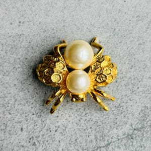 Puede incluir: Un broche de abeja de tono dorado con dos acentos de perlas blancas. El cuerpo de la abeja está texturizado con un patrón de panal.