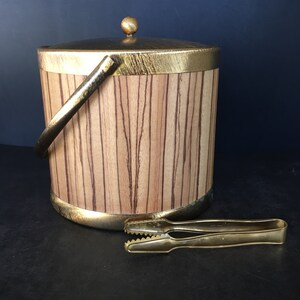 Op de afbeelding: Een vintage ijsemmer met een goudkleurige handgreep, rand en deksel. De emmer heeft een houtnerfpatroon en een kleine gouden bal bovenop. Een gouden ijstang ligt ernaast.