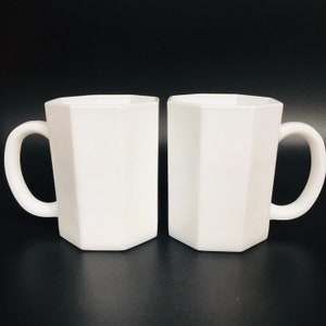May include: Two white, geometric-shaped mugs with curved handles. The mugs have a faceted design, giving them a modern look. The mugs are made of a solid white material and are set against a black background.