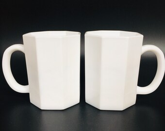 May include: Two white, geometric-shaped mugs with curved handles. The mugs have a faceted design, giving them a modern look. The mugs are made of a solid white material and are set against a black background.